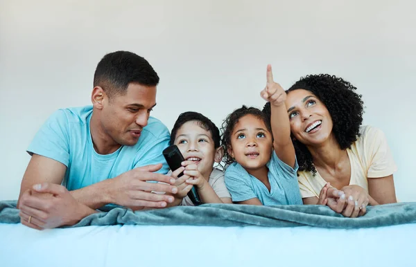Father, mom and kids on bed, watching tv and smile for comedy, funny movie or pointing in family home. Happy parents, children and watch television show with streaming, subscription or laugh together.