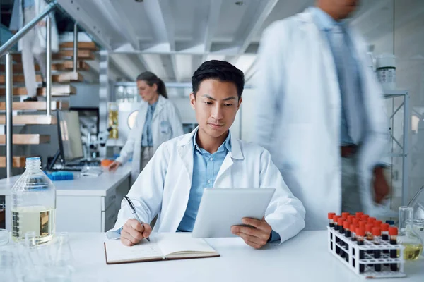 Asian man, tablet and writing in science laboratory for medical virus research, medicine and vaccine development. Busy scientist, technology and notebook paper for blood sample healthcare or wellness.
