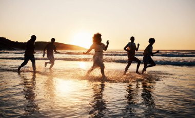 Gün batımı, plaj ve özgürlük, yaz tatilinde birlikte suda koşmak ve eğlenmek. Okyanus, siluet ve Bali 'de tatil, bağ ve seyahat kutlamaları için denizde bir grup insan..