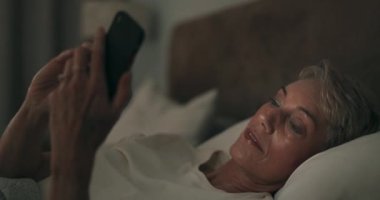 Telefon, gece ve yatakta yaşlı bir kadın ve gevşemek için mesaj okuyor. Cep telefonu, sosyal medya ve akşam yaşlı bir kadın emekliyle evinin yatak odasında tek başına yatıyordu..