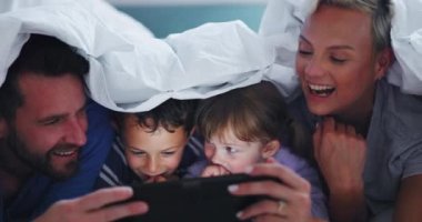 Anne, baba ve çocuklar gece yatak odasında tablet, battaniye ve oyun oynuyorlar. Aile, aileler ve çocuklar dijital teknolojiyle rahatlar, kalede online kitap medyası ile çizgi film veya hikaye anlatımı izlerler..