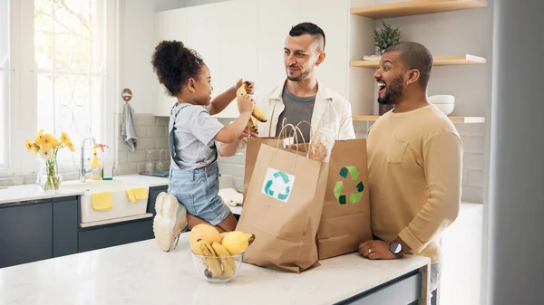 Recycling, blended family or adoption with a girl and gay couple in the home kitchen for sustainability. LGBT, eco friendly or waste with a daughter and parents unpacking bananas from a grocery bag.