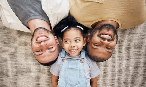 Gay dad, blended family and adoption with a girl lying together on the floor of the home for love from above. LGBT smile, children or kids and a daughter bonding with her happy parents in the house.