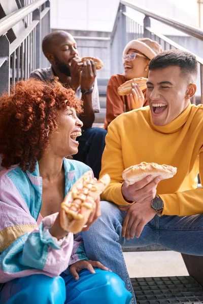 People, city and friends eating hotdog outdoor for travel, funny laugh and fun on stairs. Diversity, happiness and gen z group of men and women with food on date, adventure and freedom in urban town.