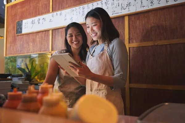 Cafe, tablet and asian women together at counter checking sales, booking or menu for small business. Social media, content marketing and startup restaurant owner with waitress scroll on digital app