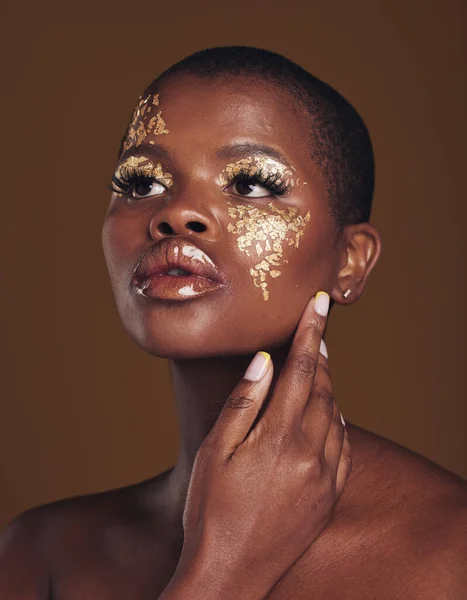 Art, aesthetic and black woman with gold makeup and brown background with glitter, paint and cosmetics. Shine, glow and African model in studio for beauty, fashion and freedom in luxury skincare
