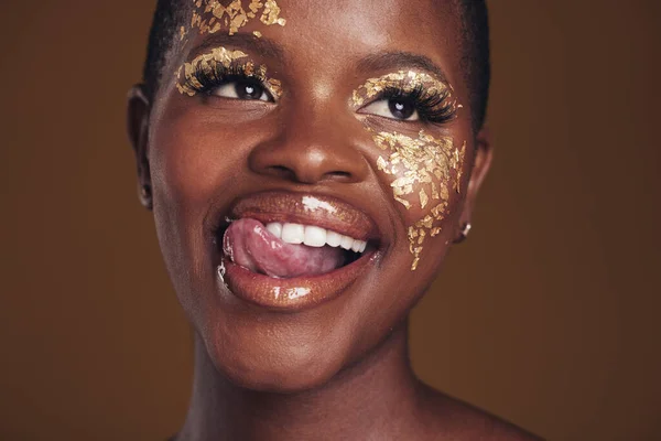 Gold, glitter and black woman beauty with makeup and tongue out in studio with sparkle cosmetics. Brown background, funny and female model with golden paint for skin glow and creative facial shine.