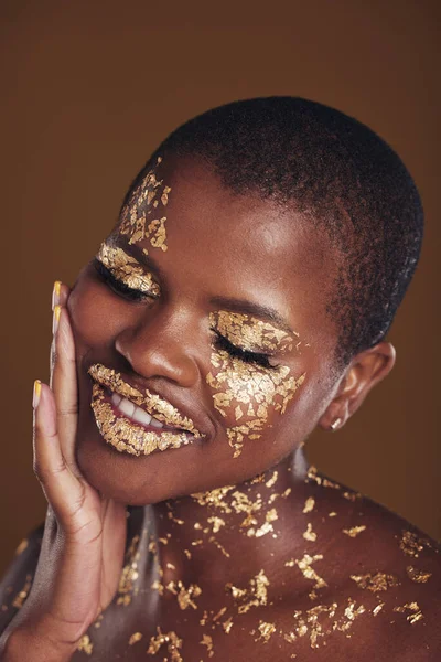Black woman, face and gold makeup with luxury glow and skincare mask with glitter. Sparkle, smile and beauty with rich and shine of cosmetics and model in studio with facial art with brown background.