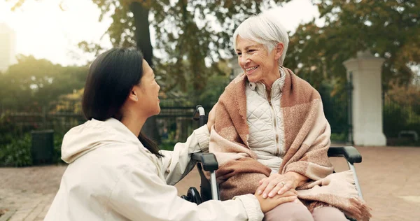 Wheelchair, park and a senior woman with a disability talking to her nurse during a walk together outdoor. Healthcare, medical and a female care chatting to an elderly patient or resident in a garden.