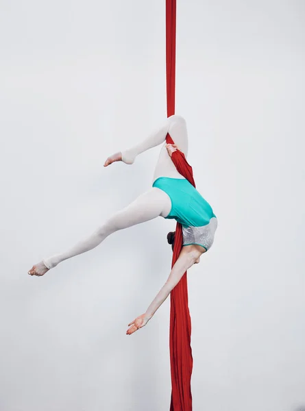 Aerial silk, acrobat and gymnastics with a woman in air for performance, sports and balance. Young athlete person or gymnast hanging on red fabric and white background with space, art and creativity.