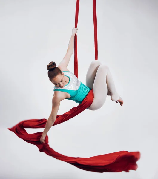 Gymnastics, aerial acrobat and sports with a woman in air for performance and balance. Young athlete person or gymnast hanging on red silk fabric and white background with space, art and creativity.