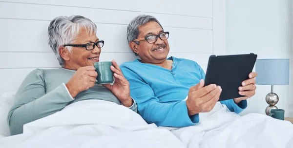 Tablet, mature couple and coffee in bed at home together on social media, streaming movie and film in the morning. Tech, man and woman drink tea in bedroom, bonding and smile reading news to relax.