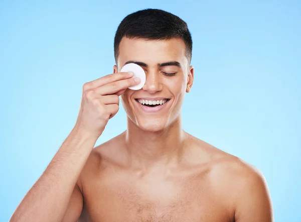 Skincare, beauty and man with cotton pad in a studio for health, wellness and self care routine. Smile, dermatology and young male model cleaning with facial cosmetic swab product on blue background.