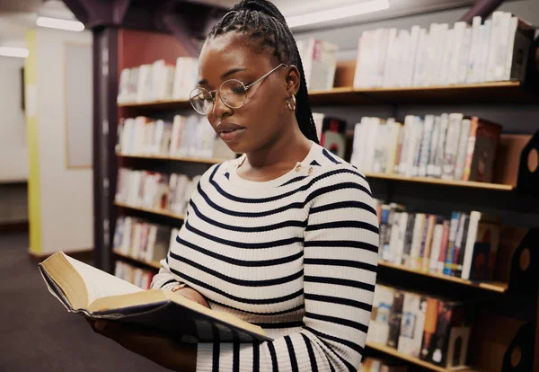 Black woman, library and books for school, student reading or education, English and language research. Young person focus by bookshelf for studying, literature catalog and free resources in glasses.