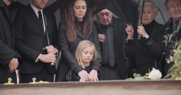 悲伤和人们在葬礼上带着雨伞 鲜花和棺材 家人带着悲伤的孩子在墓地服侍 雨中群集在墓地的棺材里 孩子在坟墓里下葬 — 图库视频影像