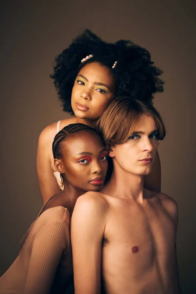 Makeup, glow and portrait of models in studio with creative, cosmetic and facial art for beauty. Queer, diversity and gen z man and women with face inclusion or skincare isolated by brown background