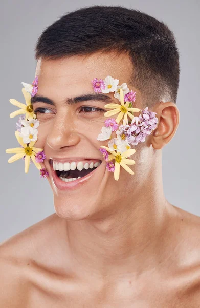 Flowers, man and skincare in studio, cosmetics and creative art on face for spring, beauty and wellness. Closeup, smile and person, facial and floral or natural glow on skin on gray background.
