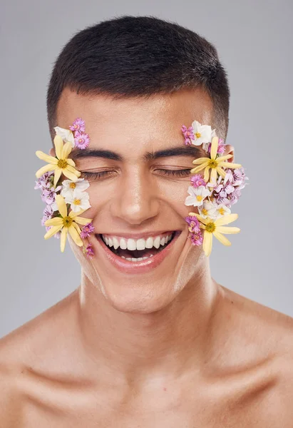 Skincare, man and flowers on face in studio, eyes closed and natural cosmetics for art, makeup product and wellness. Spring, person and closeup of floral decoration for facial on gray background.