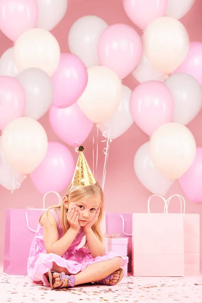Depression, sad birthday and portrait of girl on pink background for party, celebration and event in studio. Upset, emotion and unhappy, lonely and disappointed child with balloons, presents or gifts.