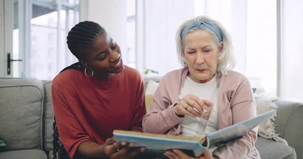 リビングルームのソファーの上に黒人女性の介護者との写真アルバム 助けられた生活と幸せなシニア おかしな 高齢者の住民が引退した家で看護師と一緒に記憶を笑う — ストック動画
