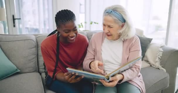 リビングルームのソファーで黒人女性の介護者と一緒に写真アルバムを助け シニアが笑った 老人ホームで看護師と一緒に記憶を見つめている笑顔 面白い 高齢者 — ストック動画
