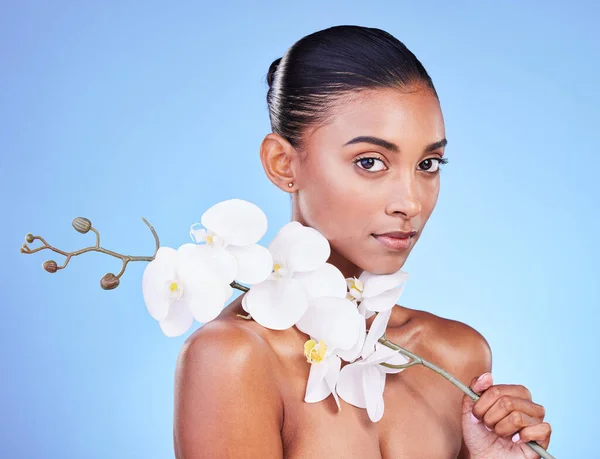Natural beauty, woman and face, flower and glow with wellness and eco friendly dermatology isolated on blue background. Sustainable skincare, orchid with collagen and cosmetics in portrait in studio.