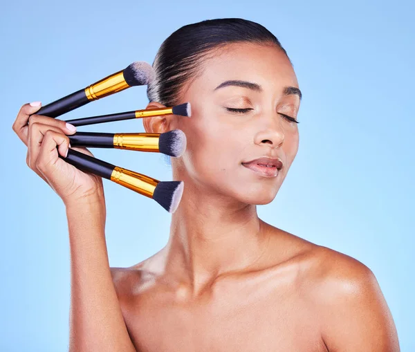Woman, makeup and face brush in studio for beauty, cosmetics and aesthetic tools on blue background. Indian model, facial dermatology and eyes closed with set of skincare products for transformation.