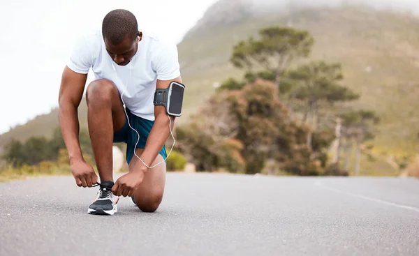 Fitness, tie or runner with shoes on a road by nature for exercise, training or outdoor workout. Sports race, black man or healthy athlete on street with footwear or headphones for streaming music.
