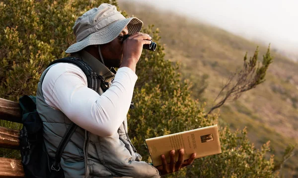 Book, binocular or man bird watching in nature on trekking adventure journey for wellness or peace. Hiking, holiday vacation or African person on bench to relax or looking to search in park for view.