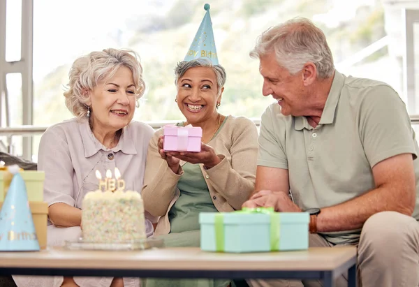 Birthday, senior friends with cake and gift for celebration in living room, smile and candles in retirement. Happiness, old people and party with dessert, surprise and elderly man with women on sofa