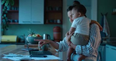 Laptop, uzaktan kumanda ya da bir anne ve bebekle akşam evde fazla mesai için finansman. Bilgisayar, aile ve iş kadını tek ebeveyn olarak bebeğiyle mutfakta birden fazla iş yapıyor..