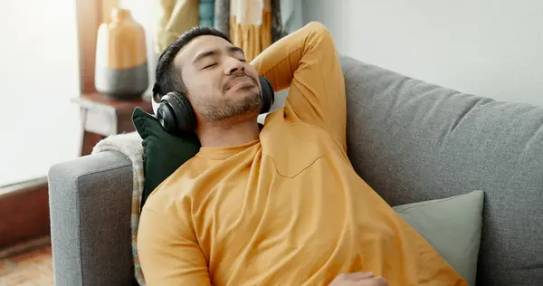 Music, headphones and asian man relax on a sofa with audio, streaming or track in his home. Earphones, radio and male with wellness app in a living room with podcast for resting, nap or day off peace.