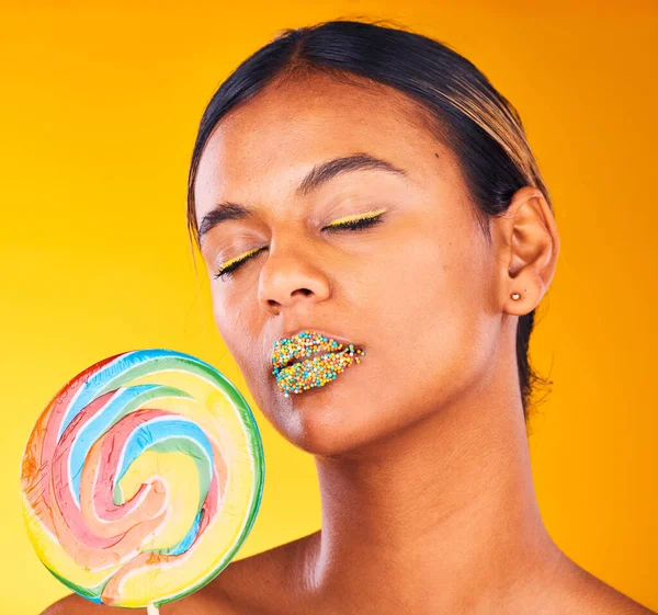 Beauty, lollipop and woman with makeup, candy and sprinkles on her lips on yellow studio background. Person, girl or model with sweet treats, aesthetic and dessert with mockup space or colorful snack.
