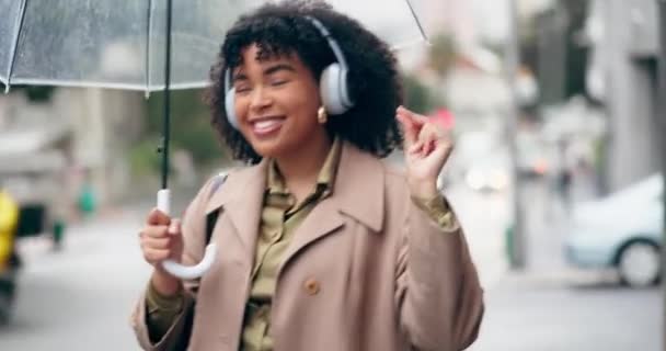 ハッピーな女性 ヘッドフォン アウトドアオーディオストリーミング用の傘で街の音楽を聴く 都会の街でサウンドトラックやダンスのためにヘッドセットで雨の中を歩く興奮した女性 — ストック動画