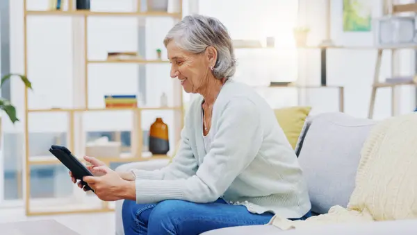 Stock image Relax, tablet and senior woman on sofa in living room scroll on social media, mobile app or the internet. Rest, online and elderly female person browsing on website with technology in lounge at home