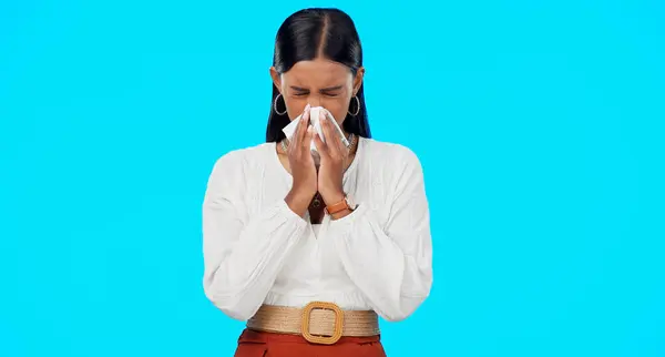 Woman, blowing nose and sick with tissue in studio for allergies, dust or pollen by blue background. Girl, sneeze and toilet paper with pain, virus and cold for healthcare, wellness and flu symptoms.