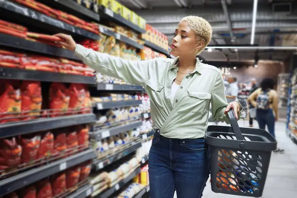 Woman, groceries and shopping basket by shelf for food price, pasta product and choice in sale or discount. African customer in convenience store or supermarket with commerce, inflation or economy.