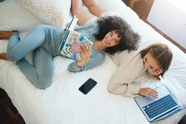 Laptop, morning and a lesbian couple relax in bed while together in their home on the weekend. Reading, computer and book with happy lgbt woman with her girlfriend in the bedroom from above for love.