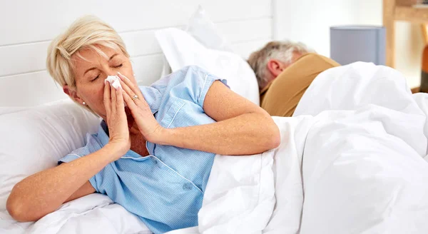 Blowing nose, couple or sick old woman in bed with husband with flu virus, cold or health problem. Sneeze, mature or senior person with tissue toilet paper, fever or allergy illness in home bedroom.