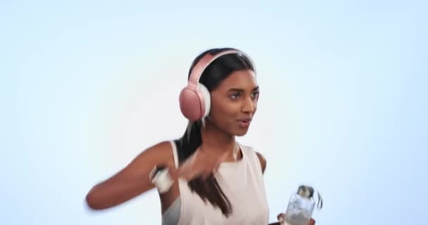 スポーツ ヘッドフォン 水分補給 エネルギー 健康のための水ボトルでスタジオで走る女性 ウェルネス フィットネス 若いインドのアスリートランナー トレーニングのためのブルーバックグラウンドで音楽を聴く — ストック動画