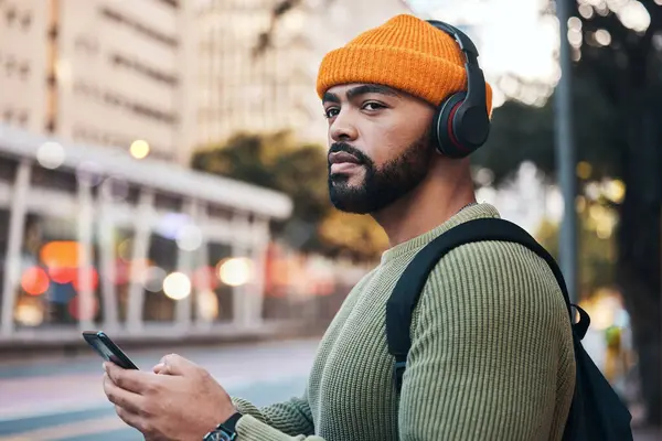 Smartphone, headphones of student and thinking in city, listening to music or sound. Phone, radio and serious man in street for streaming podcast, typing on social media and hearing audio to travel.