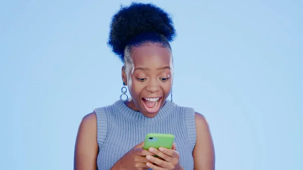 Shock, phone and black woman in a studio with good news on the internet for winning a bet. Happy, surprise and excited young African female model scroll on cellphone isolated by blue background