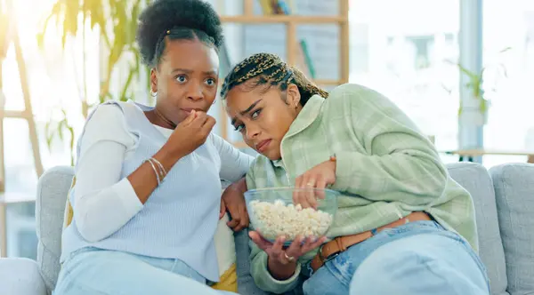 Scary, movie and friends on sofa in living room with popcorn, snack or delicious in house. Black woman, people and together by watching tv, horror or film with expression on face by sitting on couch.