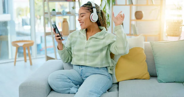 Phone, sofa and happy woman on music headphones in home living room for dancing. Mobile, radio and African person listening to podcast, audio and sound for freedom, celebration and streaming online.