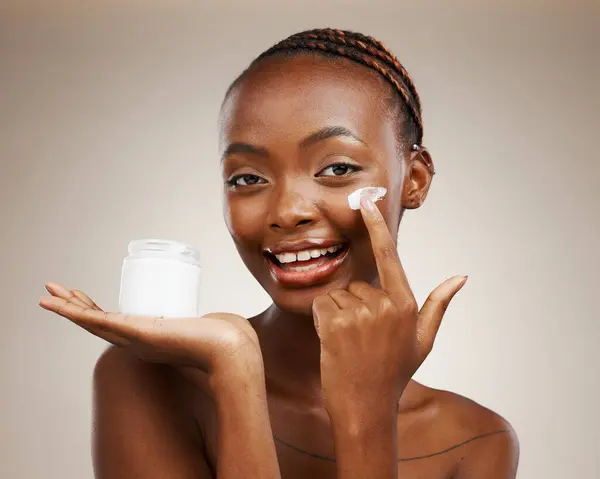 Portrait of black woman, happy or cream for wellness in studio with smile, natural cosmetics or glow. Dermatology, face or African girl model applying skincare lotion or creme on brown background.