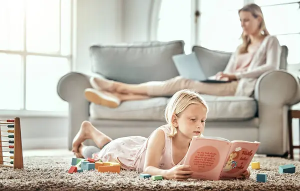 Child, floor and reading book for learning, language development and mother work from home in living room. Girl or kid with English story, education and relax on carpet with mom or family on sofa.