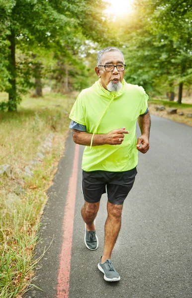 Senior man, running and road in nature, countryside or forest with exercise in retirement for cardio, health and wellness. Fitness, training and person breathing in workout, sport on run in woods.