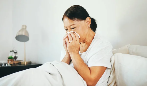 Blowing nose, bed or sick senior woman in bedroom in house with flu virus, cold or health problem. Sneeze, mature or Asian lady with tissue toilet paper, fever or allergy illness in home bedroom.