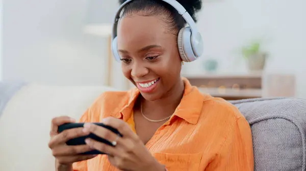 Phone games, headphones and woman in home on sofa for playing online gaming, podcast subscription or connection. Happy african person, video game or smartphone in living room for streaming multimedia.