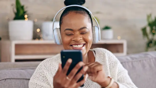 Black woman, headphones for music and smartphone, relax on couch at home and audio streaming with smile. Happiness, mobile app and using phone for radio or podcast, scroll with technology in lounge.